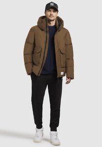 Hnedá zateplená bunda s kapucňou, dvoma prednými vreckami a zipsom. V kombinácii s čiernymi teplákmi a bielymi teniskami.