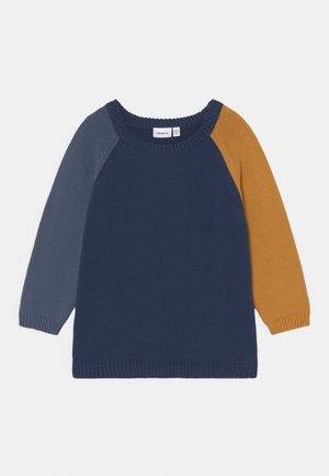 Długi sweter z dzianiny z granatowym tułowiem, jednym szarym rękawem i jednym musztardowożółtym rękawem, z okrągłym dekoltem.