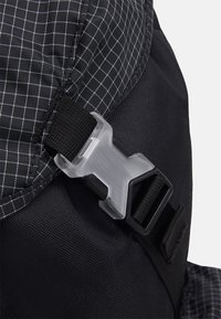 Svart ryggsäck med ett rutmönster, utrustad med ett klart plastspänne och svarta remmar. Tyget verkar strukturerat och hållbart.