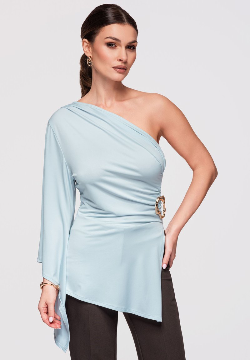 Femme aux cheveux bruns en queue de cheval basse porte un haut bleu clair asymétrique avec une boucle dorée et un pantalon noir sur un fond uni.
