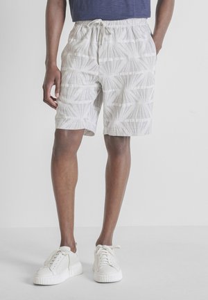 Hombre con pantalones cortos blancos con cordón y estampado, camisa azul marino y zapatillas blancas, de pie frente a un fondo gris liso.