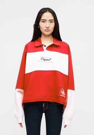 Jeune personne aux longs cheveux foncés portant un pull de style rugby Dsquared2 rouge et blanc et un jean foncé, debout devant un fond uni.