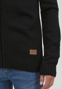 Svart strikket cardigan med ribbestrikkede mansjetter og nederkant. Har et brunt lærmerke med "11 PROJECT" på forsiden. Strukturert stoff.