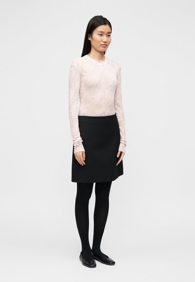 Haut à manches longues rose clair avec un motif subtil, associé à une jupe noire, des collants noirs et des chaussures plates noires.