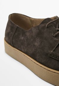 Brown suede lace-up shoe with a rounded toe, flat tan rubber sole, and simple stitching details along the edge.