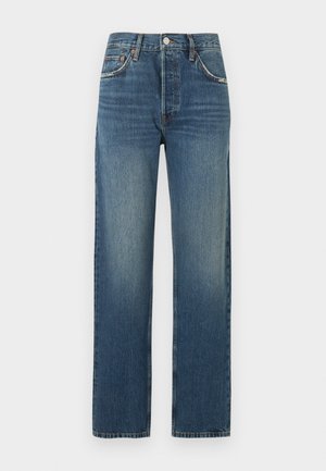 Vrouw staand met hand in de zak, draagend een beige top met lange mouwen, hoog getailleerde blauwe spijkerbroek en zwarte sandalen met open teen en hak, op een effen achtergrond.