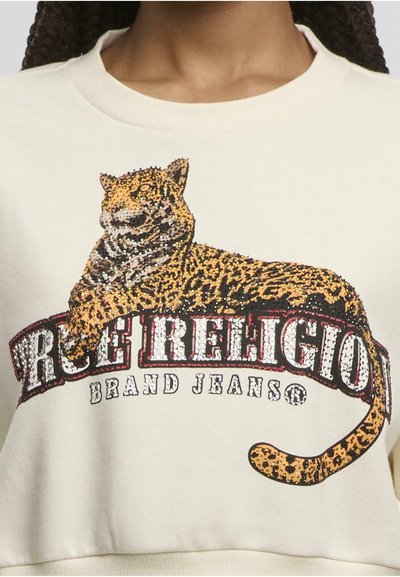 Graphique de léopard avec des détails scintillants au-dessus du texte "True Religion Brand Jeans" sur un sweat-shirt de couleur crème porté par une personne.