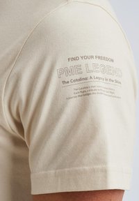 Bež majica iz bombaža s tiskanim dizajnom na lijevom ramenu koji uključuje tekst "PME Legend" i "Find Your Freedom." Tekstura je glatka.