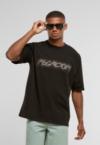Sort bomulds oversized t-shirt med et gråt grafisk logo. Modellen har sorte solbriller på, og mintgrønne denim bukser er synlige.