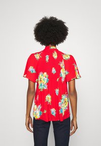 Röd blommig blus i lätt tyg, med korta ärmar, plisserad textur och ljusfärgade blommönster.