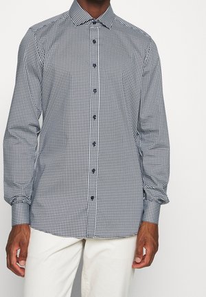 Man wearing a black and white checkered long-sleeve button-up shirt with white pants, standing against a plain background.