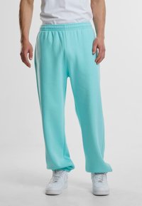 Turkise joggebukser laget av myk stoff, med elastisk midje, avslappet passform og samlet mansjetter ved anklene. Brukt med hvite sneakers.