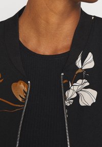 Chaqueta bomber negra con bordados florales en blanco y marrón, con un cuello acanalado y un cierre de cremallera plateado en la parte delantera.