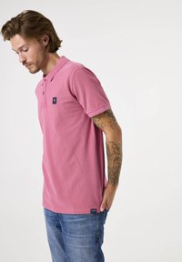 Camisa polo rosa con cuello clásico, mangas cortas y un parche de logo. Combinada con jeans de mezclilla azul, de corte relajado y con un tatuaje visible.