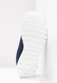 Semelle texturée blanche d'une chaussure de sport bleu marine reposant verticalement contre un mur d'angle blanc sur une surface blanche.