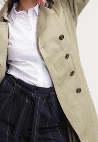 Beige dobbeltradet frakke med store brune knapper, parret med en hvid skjorte med krave og mørke stribede bukser med en bundet talje.