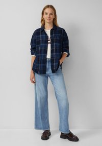 Marine ruit flanellen overhemd, dichtgeknoopt, over een wit graphic T-shirt. Lichtblauwe wijde denim jeans. Stevige bruine schoenen. Glad textuur.