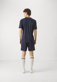 Male athlete standing facing away, wearing navy blue sportswear, white knee-high socks with black stripes, and white cleats on a white background.