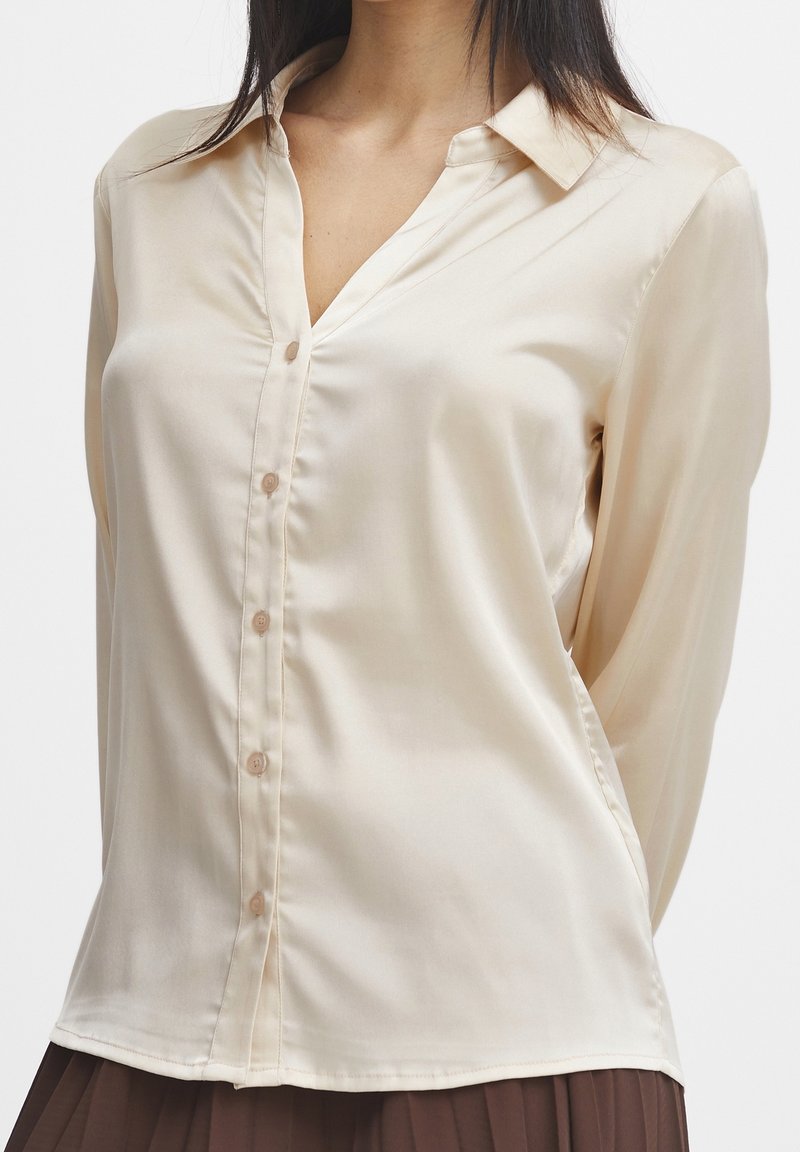 Blouse beige clair à manches longues avec un devant à boutons, col cranté et texture satin lisse. Présente un éclat subtil et une coupe ajustée.