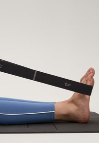 Črn raztegljiv trak držimo ob stopalu, z natančno sivo dotikom. Vidne so modre legice z belim robom, postavljene na teksturirano črno podlogo.