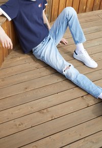 Marineblaues Kurzarmhemd mit weißen Akzenten, hellblaue ausgewaschene Jeans und weiße Sneakers mit blauen Details, auf Holzdecke sitzend.