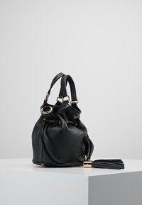 Sac seau en cuir noir avec quincaillerie en tonalité dorée, fermeture à cordon, détail de frange sur le côté et finition douce et texturée.