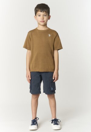 Ragazzo in piedi che indossa una maglietta marrone a maniche corte, pantaloncini cargo blu navy e scarpe da ginnastica bianche con lacci neri su sfondo chiaro uniforme.