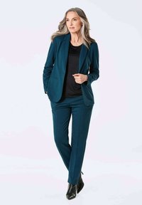 Een teal blazer en bijpassende tapered broek van soepel materiaal, gecombineerd met een zwarte top. De outfit heeft een getailleerde pasvorm en strakke lijnen.