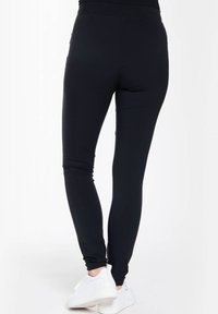 Zwarte legging van rekbaar materiaal met een gladde textuur, voorzien van een hoge tailleband en een enkellang ontwerp, gecombineerd met witte sneakers.