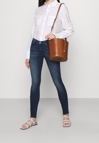 Camisa blanca de botones, jeans skinnies oscuros, bolso de cuero marrón tipo cubo y sandalias blancas de tiras. Diseño simple con características funcionales.