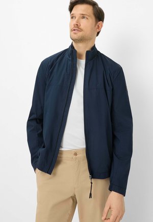 Man draagt een marineblauw jasje met rits over een wit hemd en beige broek, staat met handen in de zakken, kijkt omhoog naar rechts.