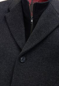 Cappotto di lana grigio antracite con colletto a revers, chiusura frontale con bottoni e uno strato interno con zip rivestito da una fodera rossa decorata.