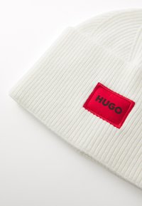 White ribbed knit beanie featuring a red rectangular patch with the black text "HUGO" in the center. Soft texture and close-fitting design.