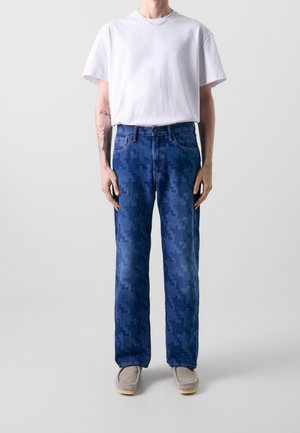 Person trägt ein schlichtes weißes T-Shirt, blaue Jeans mit Pixelmuster, beigen Schuhe und eine dünne Kettenhalskette, steht vor einem hellgrauen Hintergrund.