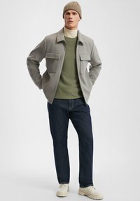 Graue Wolljacke mit zwei Fronttaschen, darunter ein grüner Pullover, blaue Jeans und beigefarbene Schuhe. Modell trägt eine gerippte beiger Mütze.