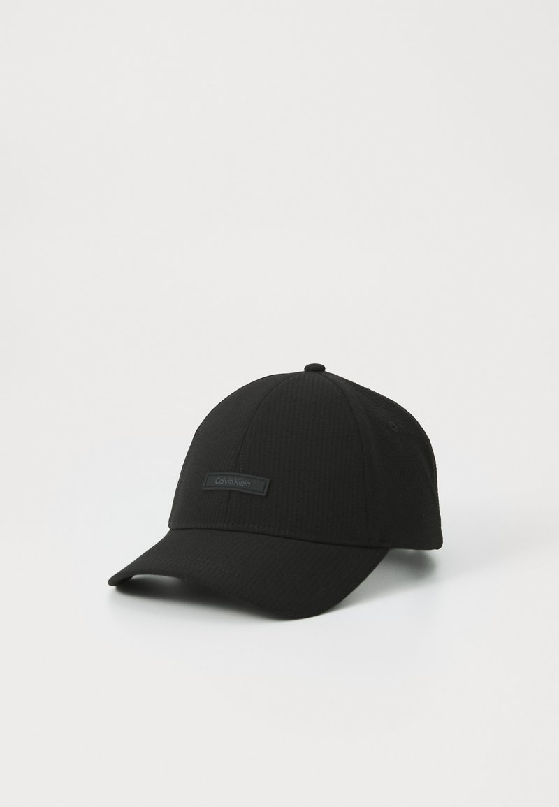 Casquette de baseball noire en tissu texturé avec une couronne arrondie et une visière plate. Présente un accent de logo rectangulaire à l'avant.