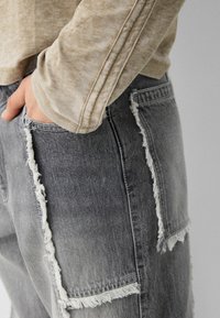 Světlé šedé rifle z vybledlé džínoviny s ošoupanými záplatami a výraznými bočními švy, kombinované s béžovým tričkem s dlouhým rukávem.