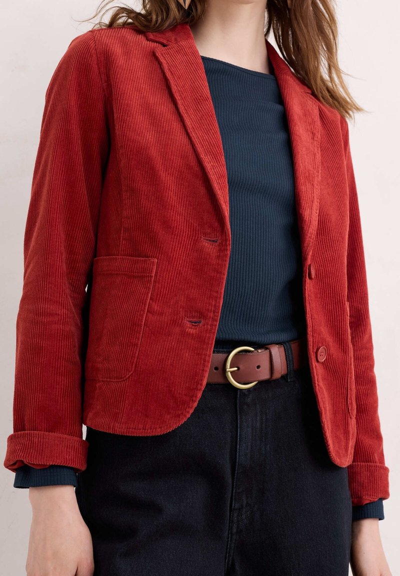 Femme portant un blazer en velours côtelé rouge rouille sur un top côtelé bleu marine, un pantalon noir taille haute et une ceinture marron avec une boucle dorée.