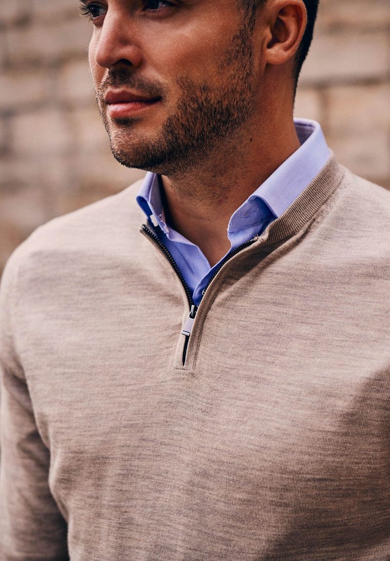 Beiger Zip-Hals-Pullover mit strukturierter Oberfläche, über einem hellblauen Hemd mit Kragen getragen. Eine Nahaufnahme hebt die Stoffdetails und Nähte hervor.