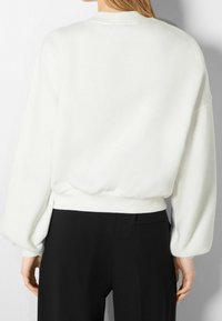 Biała bluza z balonowymi rękawami, prążkowanymi mankietami i dołem. Miękka tkanina, luźny krój, noszona na czarnych spodniach z boczną kieszenią.