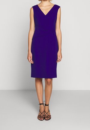 Femme portant une robe violette sans manches, longueur genou, avec un col en V et des sandales noires à talons hauts à brides, debout devant un fond uni.