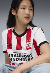 Ung person iförd röd och vit randig PSV Eindhoven fotbollströja med sponsor- och Puma-logotyper, sittande med benen i kors.