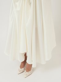 Λευκό σατέν φόρεμα με ρέουσα σχεδίαση και γυαλιστερή επιφάνεια, συνδυασμένο με λευκές γόβες με μυτερή μύτη που διαθέτουν διακριτικά λουλουδάτα μοτίβα.