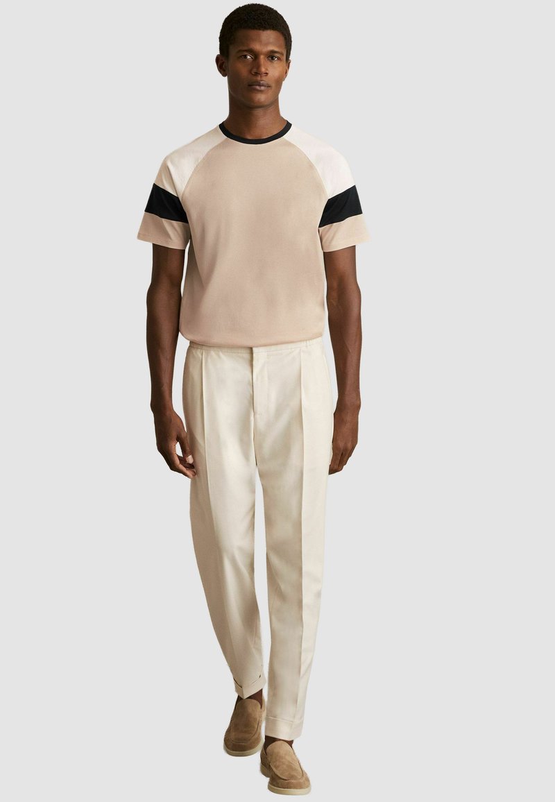 Bežs īsām piedurknēm T-krekls ar melnbaltiem akcentiem, apvienots ar piegrieztiem krēma biksēm un brūniem apaviem, ar brīvu piegriezuma dizainu.