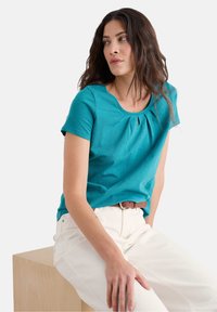 Tyrkysový krátký rukáv top s kulatým výstřihem a malým detaily ve tvaru klíče. V kombinaci s bílými kalhotami a hnědým páskem, sedící na světlém dřevěném bloku.