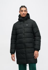 Νέος με πλεξούδες φοράει ένα μαύρο padded μπουφάν με κουκούλα και φερμουάρ και πράσινα παντελόνια, στέκεται μπροστά σε λευκό φόντο.