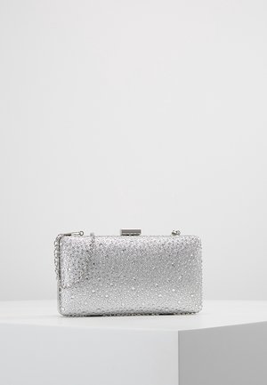 Pochette rectangulaire argentée avec surface texturée en strass et une fine bandoulière en chaîne, posée sur un piédestal blanc.