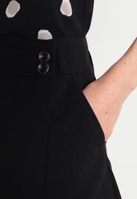 Pantaloni neri su misura con due bottoni, tasche laterali e texture liscia. Il tessuto appare strutturato e leggero.