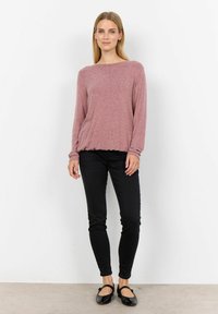 Lange mouwen, lichte roze trui met een wijde halslijn, gecombineerd met zwarte skinny jeans en zwarte flats met strikaccenten.