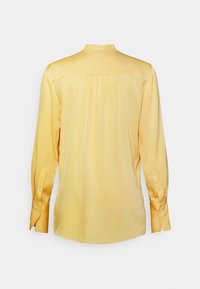 Camicia di seta gialla con plissè sul retro, maniche lunghe con polsini a bottone e colletto alto. Tessuto liscio e colore solido senza motivi.